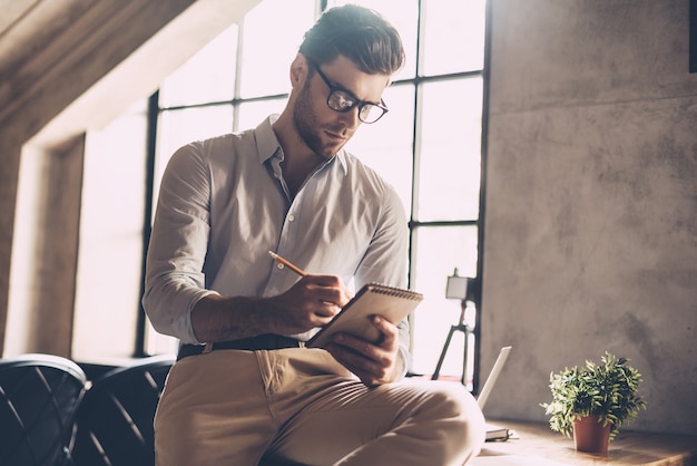 Juste inspiré. Jeune homme confiant dans des vêtements décontractés intelligents écrivant quelque chose dans son cahier tout en se penchant au bureau au bureau