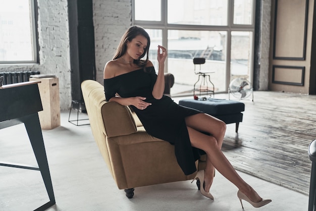 Juste parfait. Jolie jeune femme en robe noire élégante avec une fente profonde relaxante assise sur le canapé