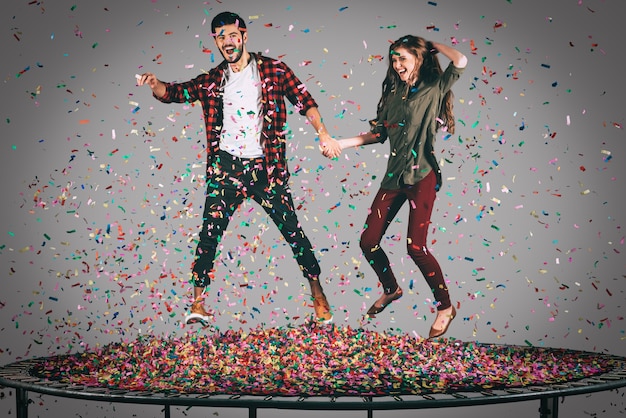 Juste pour le fun. Photo en l'air d'un beau jeune couple joyeux se tenant la main tout en sautant sur un trampoline avec des confettis tout autour d'eux