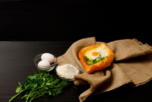 Khachapuri avec oeufs sur un sac, farine, oeufs et persil sur tableau noir, fond