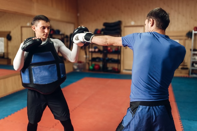 Kickboxer masculin dans les gants pratiquant le coup de main avec un entraîneur personnel en coussinets, entraînement en salle de sport. Boxeur en formation, pratique du kickboxing