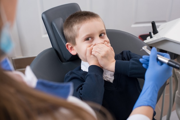 Kid effrayé par les dentistes couvre sa bouche et le dentiste dans des gants tenant dans la mèche dentaire