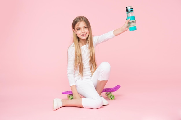 Kid fille soins hydratation du corps. Loisirs actifs et équilibre hydrique. Un enfant actif et en bonne santé boit de l'eau. Rester hydraté. Le visage heureux de la fille tient avec une bouteille d'eau tout en s'asseyant sur fond rose de planche de penny.