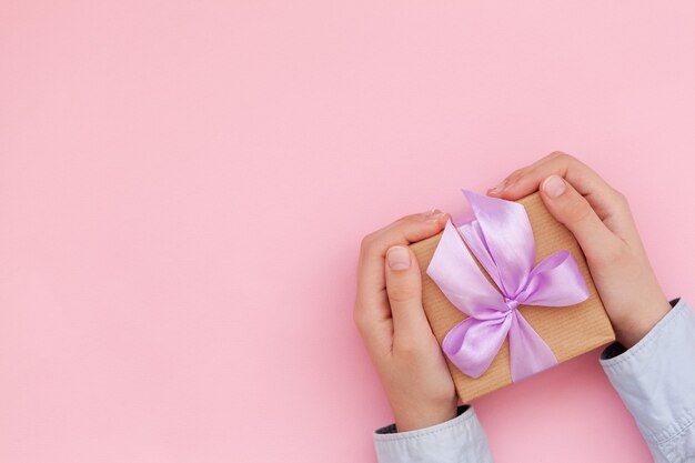 Kid's hands holding boîte-cadeau enveloppé dans du papier kraft et attaché avec un arc sur le mur rose.