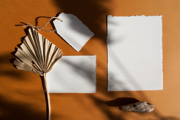 Kit de cartes d'invitation de mariage en papier blanc vierge affiche bricolage et étiquette avec feuille de palmier séchée sur table en terre cuite texturée. Modèle moderne élégant pour l'identité de marque. Mise à plat, vue de dessus