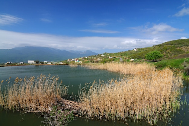 Lac aux eaux boueuses et roseaux en croissance, ciel bleu et petit village parmi les montagnes