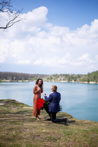 Lac dans la forêt : un homme fait une surprise à sa petite amie, il lui met une bague au doigt, une déclaration d'amour et une offre de mariage. Fille avec un sourire sur son visage et un bouquet de roses rouges