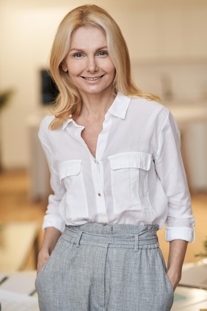 Lady boss femme mature réussie avec des cheveux blonds souriant à la caméra en se tenant debout à l'intérieur