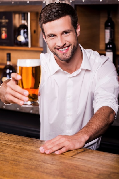 Laissez-moi étancher votre soif ! Beau jeune homme barman en chemise blanche étirant le verre avec de la bière et souriant tout en se tenant au comptoir du bar