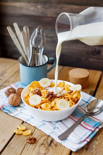 Le lait d&#39;une bouteille coule dans le bol avec de délicieux flocons de maïs aux noix et à la banane. Fond en bois rustique avec une serviette à carreaux. Collation santé au petit déjeuner croustillant.