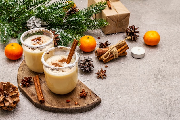 Lait de poule à la cannelle et à la muscade pour les vacances de Noël et d'hiver. Boisson maison dans des verres à bord épicé. Mandarines, bougies, cadeau.