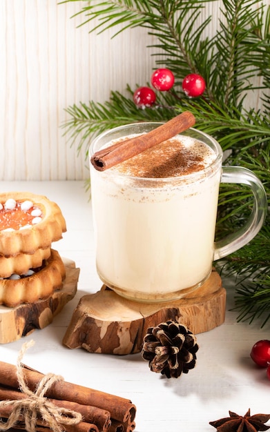 Lait de poule crémeux de Noël à la cannelle, pile de biscuits au gingembre sur une table en bois blanc avec des branches de sapin.