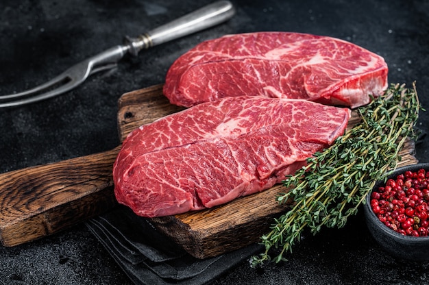 Lame supérieure crue fraîche ou steaks de viande de bœuf en fer plat sur une planche à découper de boucher. Fond noir. Vue de dessus.