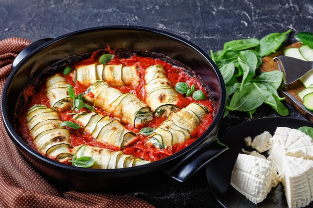 Lanières de courgettes roulées avec une garniture de ricotta mélangée à de jeunes épinards, feuilles de basilic frais cuites au four dans une sauce tomate servi sur un plat de cuisson noir rond sur une table en béton foncé, gros plan