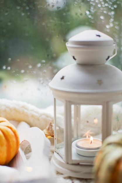 Lanterne blanche avec bougie allumée et décorations d'automne. Citrouilles orange, plateau en bois et lanterne sur un pull en tricot blanc sur le rebord de la fenêtre