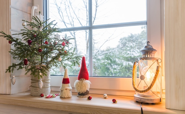 Lanterne de Noël, gnome de Noël, sapin de Noël et décor rouge sur la fenêtre d'une maison en bois donnant sur le jardin d'hiver. Composition de Noël