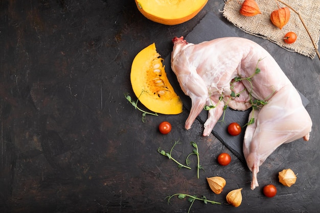 Lapin cru entier avec citrouille, tomates, pousses de pois sur fond de béton noir et textile en lin. Vue de dessus, mise à plat, espace de copie.