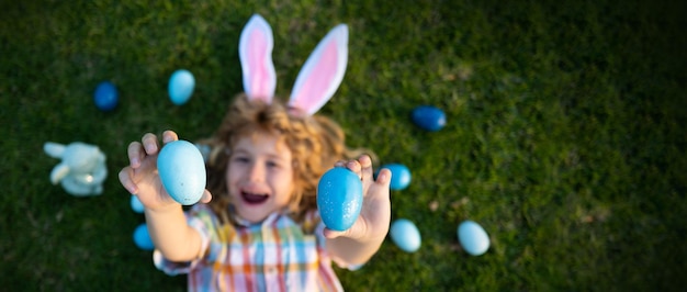 Lapin enfant mignon porter des oreilles de lapin dans le jardin Joyeux jour de Pâques Lapin garçon drôle portrait en plein air Vue de dessus visage drôle d'enfant Bannière photo horizontale pour la conception d'en-tête de site Web