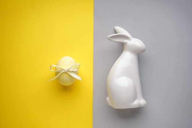 Lapin de Pâques en céramique avec oeuf de pâques jaune avec ruban sur une surface jaune-gris, mise à plat.