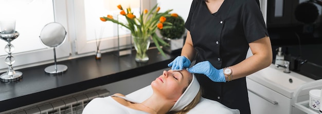 Large photo en intérieur d'une cosméticienne méconnaissable en uniforme noir et gants de protection bleus utilisant des échanges cosmétiques sur le visage de sa cliente caucasienne massage SPA