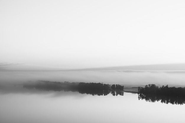 Une large rivière mystique coule le long de la rive diagonale avec une silhouette d'arbres et un épais brouillard en niveaux de gris. Ambiance laiteuse du matin. Paysage monochrome minimaliste de fond de nature majestueuse