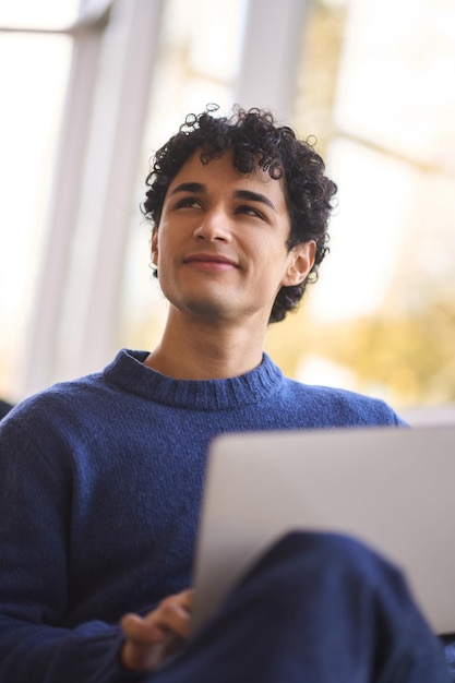 Un latin souriant regardant rêveusement de côté le raisonnement sur son projet tout en travaillant en ligne sur un ordinateur portable