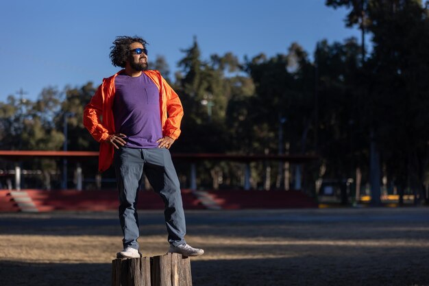 Latino avec la barbe et les cheveux bouclés sur un tronc dans un parc posant fièrement