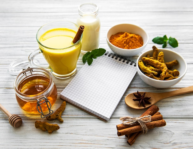 Latte au curcuma doré dans un verre, épices et livre de recettes sur fond de bois blanc