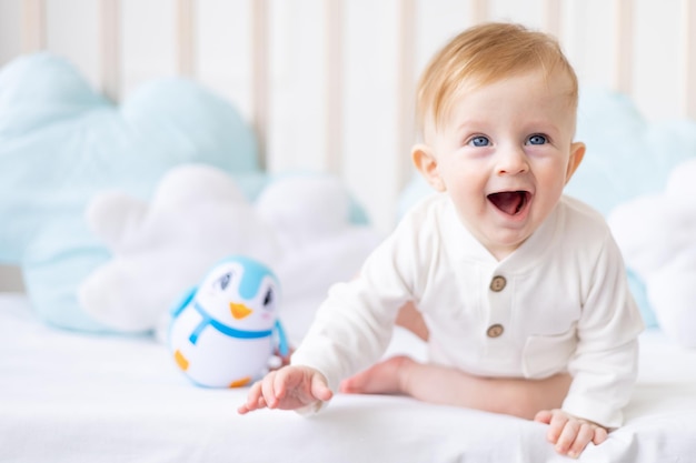 Laughing baby 6 mois garçon blond dans un berceau dans une chambre lumineuse dans un body en coton blanc avec un jouet bleu le concept de produits pour enfants