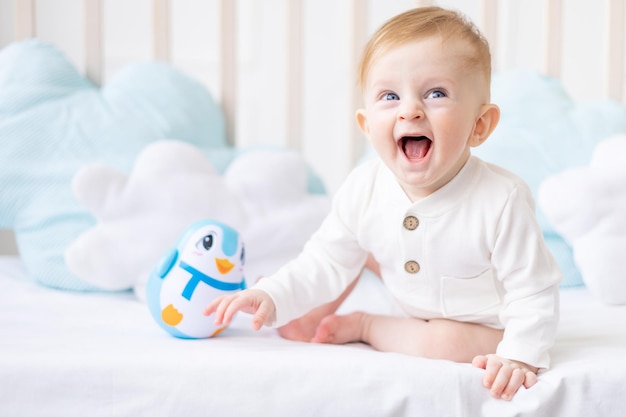 Laughing baby 6 mois garçon blond dans un berceau dans une chambre lumineuse dans un body en coton blanc avec un jouet bleu le concept de produits pour enfants