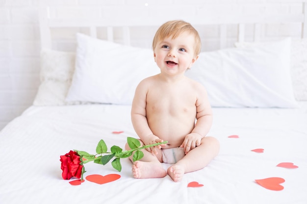 Laughing baby boy blonde est assise sur le lit en couches et avec une rose rouge à la maison et souriant parmi les cœurs le concept de la Saint-Valentin ou de la fête des mères