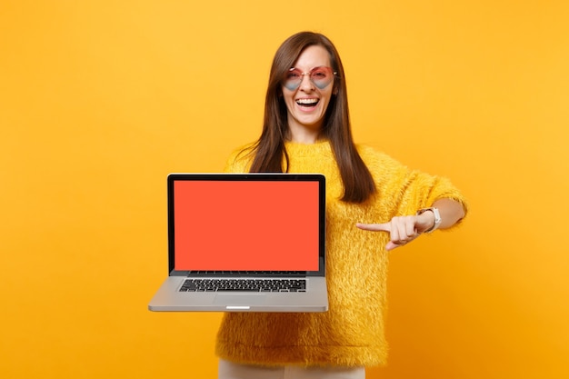 Laughing woman in heart verres pointant l'index sur ordinateur portable pc avec écran vide noir blanc isolé sur fond jaune vif. Les gens émotions sincères, mode de vie. Espace publicitaire.