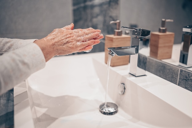 Lavage à la main mousse de savon liquide en frottant les poignets étape de lavage des mains femme senior rincer dans l'eau à l'évier du robinet de salle de bains.