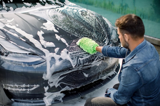 Lavage De Voiture En Plein Air En Libre-service Vue En Angle Arrière Du Beau Jeune Homme De Race Blanche Lavant Une Voiture électrique Grise De Luxe Avec Une Brosse Verte Et De La Mousse