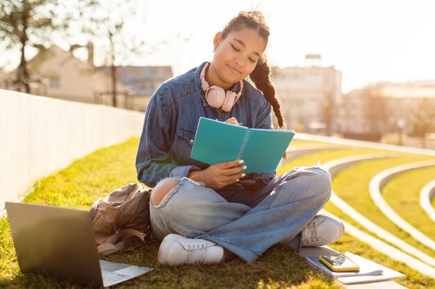 Leçon à distance étudiante métisse étudiant à l'extérieur avec un ordinateur portable assis dans le parc et prenant