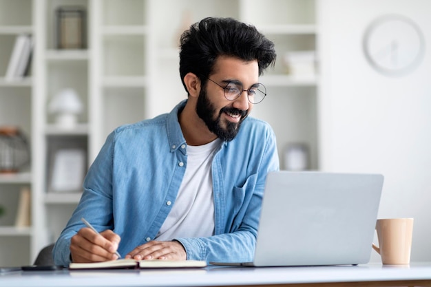 Leçon en ligne jeune homme indien souriant utilisant un ordinateur portable et prenant des notes