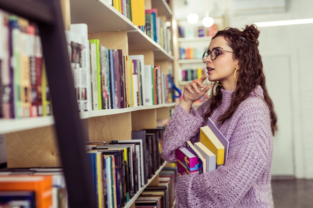 Lecteur collectant activement. Dame positive réfléchie choisissant entre deux livres tout en restant devant des étagères