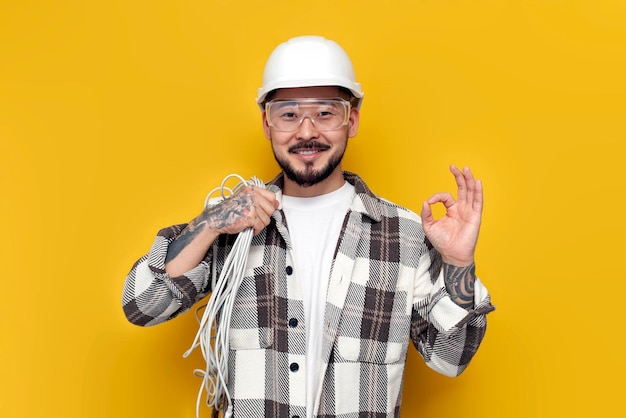 Électricien asiatique mâle en casque et lunettes tient le câble internet et montre ok avec sa main