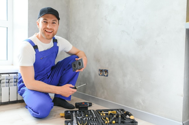 Électricien professionnel souriant en uniforme remplaçant une prise dans un appartement après des travaux de rénovation