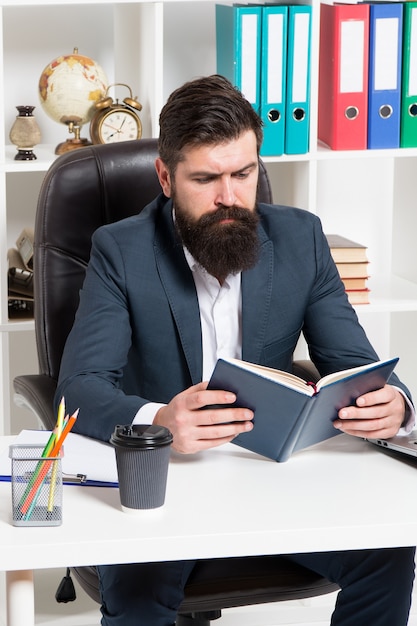 Lecture d'un livre d'affaires d'autorité. L'homme barbu patron hipster s'asseoir fauteuil bureau intérieur lire livre. Patron au travail. Guy travail de style d'entreprise de vêtements formels. Littérature commerciale. Le patron a lu le livre.