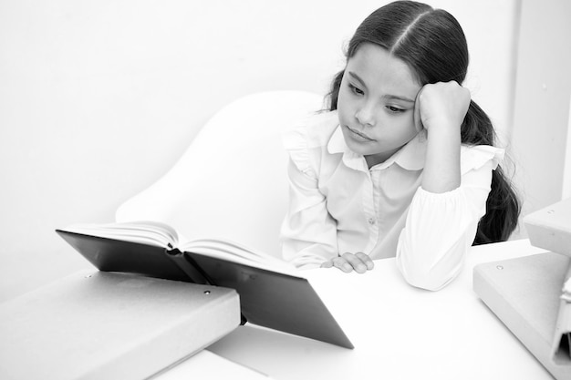 Lecture d'un livre ennuyeux Petite fille lit un livre tout en s'asseyant sur une table fond blanc Écolière étudiant et lisant un livre