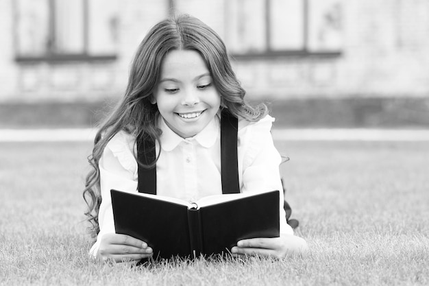 Lecture parascolaire Mignon petit enfant lisant un livre à l'extérieur Adorable petite fille apprend à lire Uniforme scolaire d'écolière allongé sur la pelouse avec son livre préféré Concept d'étude Éducation de base
