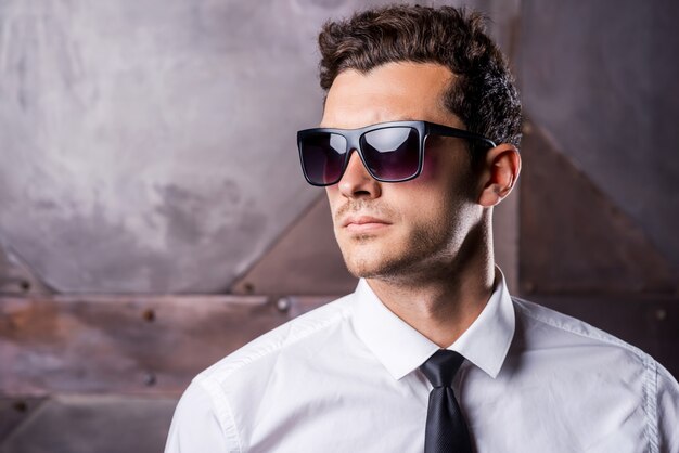 Élégant beau. Beau jeune homme en tenue de soirée et lunettes de soleil en détournant les yeux en se tenant debout sur fond de métal