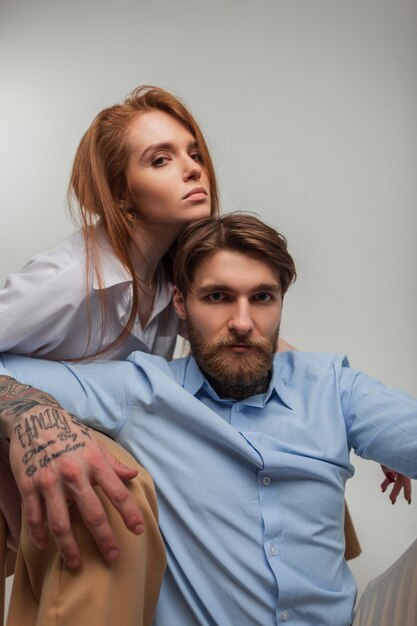 Élégant beau jeune couple dans des vêtements élégants à la mode sont assis sur un fond blanc Beau modèle de femme rousse et bel homme hipster avec tatouage et barbe