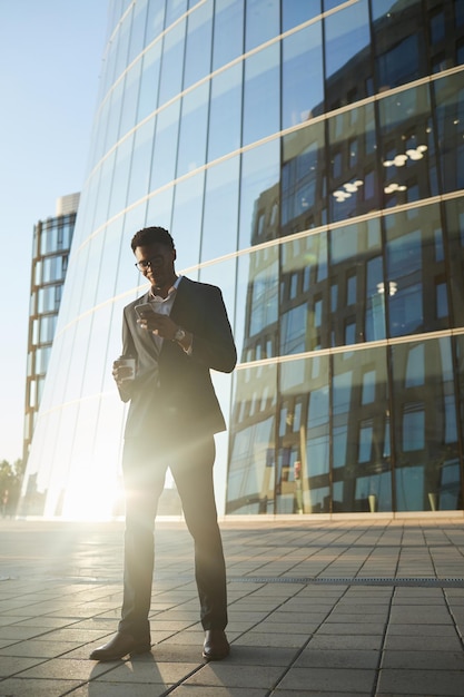 Élégant beau jeune homme d'affaires noir à lunettes utilisant un smartphone et buvant du café en mouvement