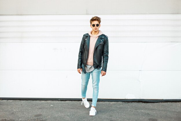 Élégant beau jeune homme avec des lunettes de soleil dans une veste en cuir noir, un jean à la mode bleu et des baskets à la mode blanches près d'un mur en métal blanc