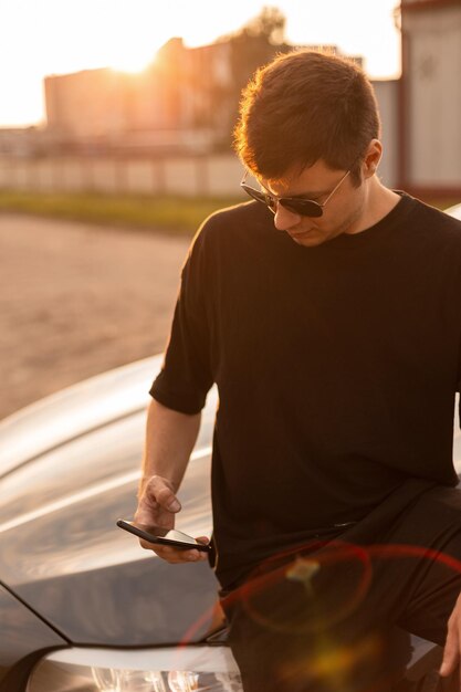 Élégant et beau mec hipster avec des lunettes de soleil cool et un t-shirt noir à la mode regarde son téléphone et est assis près de sa voiture au coucher du soleil