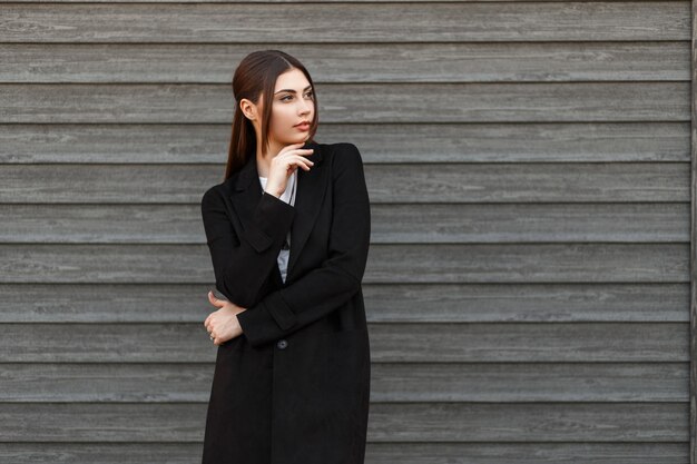 Élégant beau modèle femme en manteau noir posant près de maison en bois vintage