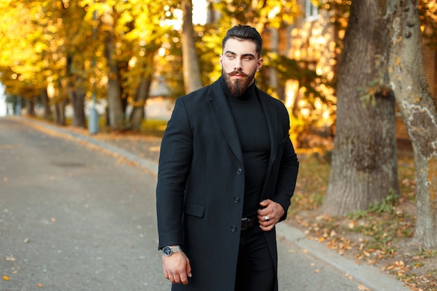 Élégant bel homme dans un manteau noir marchant à l'extérieur sur une journée ensoleillée d'automne