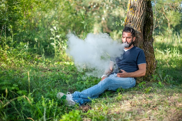 Élégant homme vape barbu fumant un appareil à fumée électronique dans la forêt. Vape.